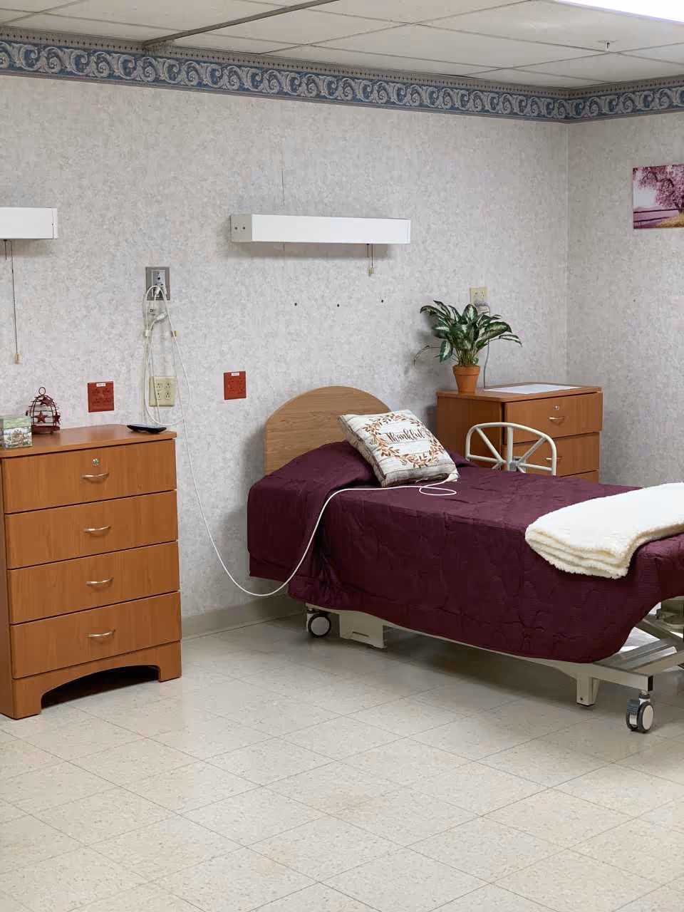 A simple nursing facility bedroom with a single bed covered in a dark purple quilt and a white blanket folded at the foot. There is a pillow with the word 'Thankful' on it. The bed has wheels and is positioned between two wooden dressers. A potted plant sits on the dresser to the right. The walls are light-colored with a decorative blue and white border near the ceiling. Medical equipment outlets and cords are visible on the wall behind the bed.