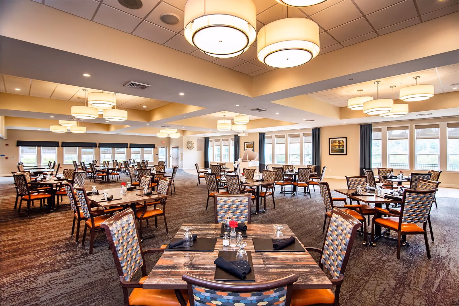A spacious dining room with multiple tables and chairs arranged neatly. Each table is set with black placemats, upside-down glasses, black napkins, and salt and pepper shakers. The room has large windows with blue curtains allowing natural light to enter. The ceiling features modern circular light fixtures, and the carpet has a textured pattern. The walls are painted a light beige color with framed artwork.