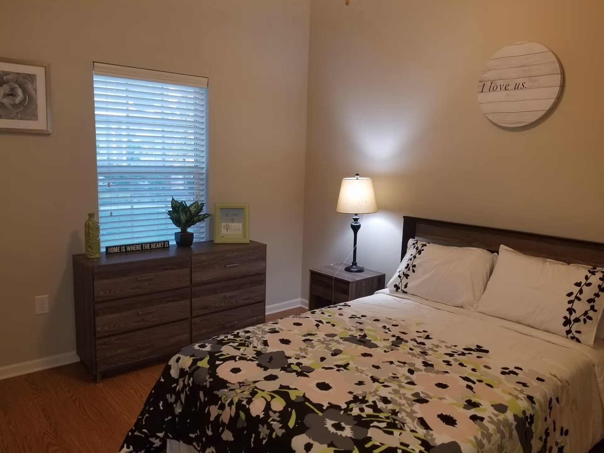 A cozy bedroom with a bed covered in a floral patterned comforter and white pillows. Next to the bed is a nightstand with a lit lamp. Across from the bed is a wooden dresser with a plant, a framed sign, and a decorative item on top. A window with blinds is above the dresser. On the wall above the bed is a round wooden sign that reads 'I love us.'