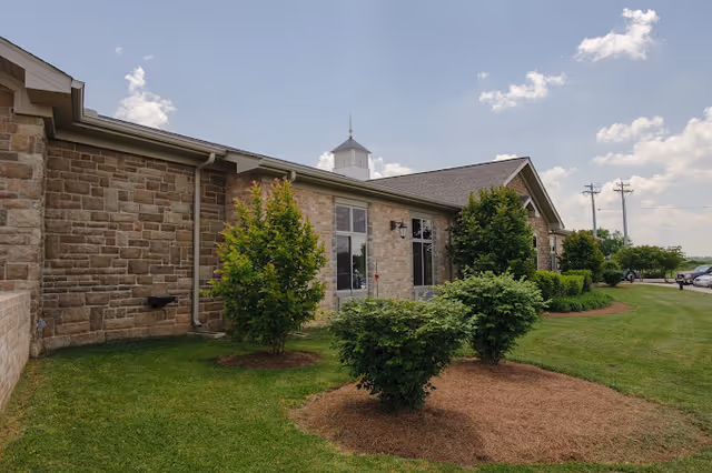 Stone-faced senior living facility exterior with windows, landscaped shrubs and a grassy lawn under a partly cloudy sky.