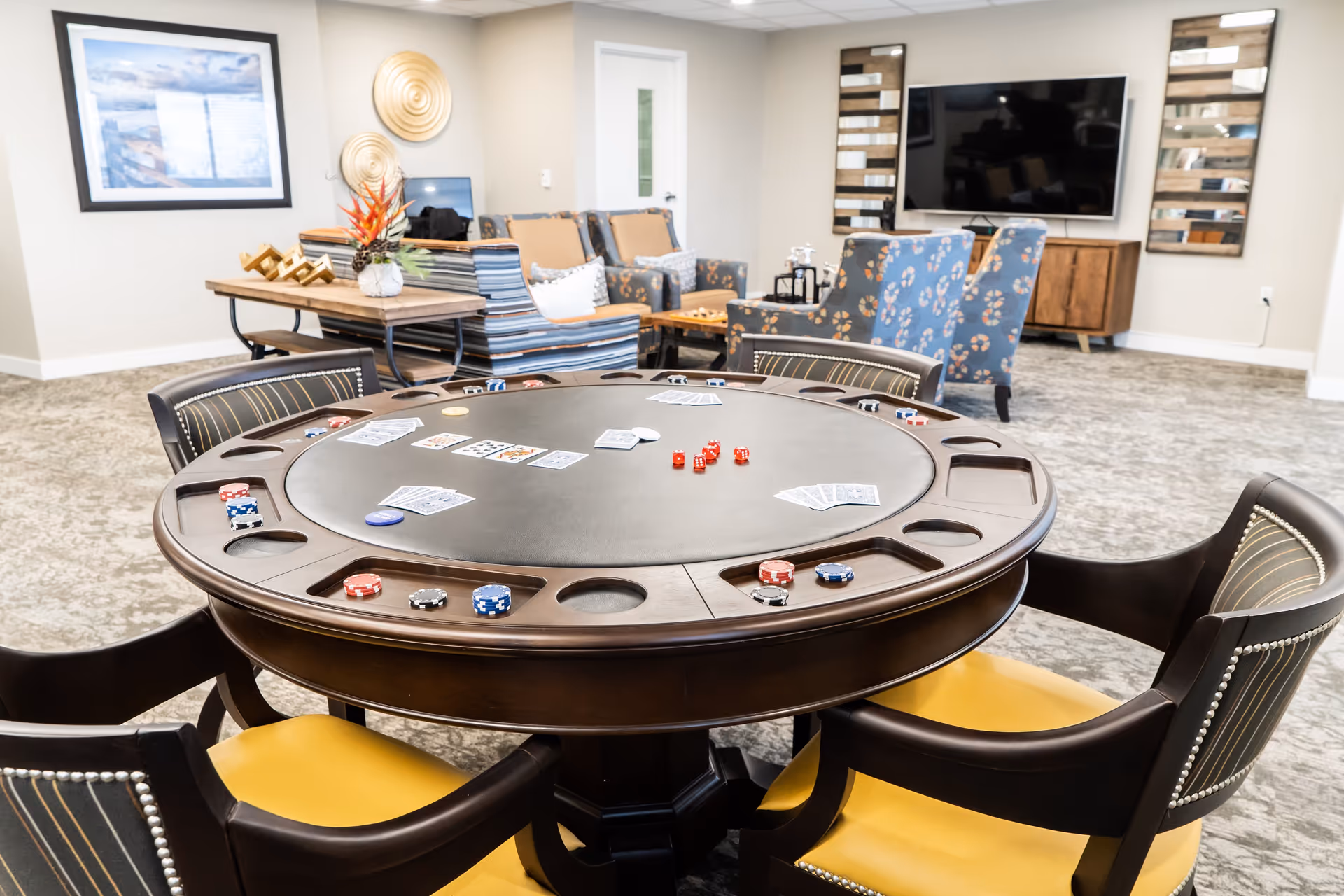 A cozy game room featuring a round poker table with playing cards, poker chips, and red dice on it. Surrounding the table are chairs with dark wood frames and yellow cushions. In the background, there are comfortable armchairs with patterned upholstery, a wooden console with a large flat-screen TV mounted on the wall, decorative wall art, and a table with a floral arrangement.