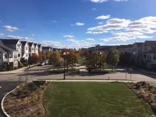 View of a residential community with multiple two- and three-story buildings on either side of a street. There is a landscaped median with trees and street lamps in the center, and a grassy area with some plants in the foreground under a partly cloudy blue sky.