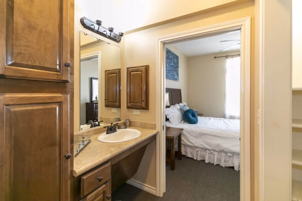 View of a bathroom vanity with a sink, mirror, and wooden cabinets. Through an open doorway, a bedroom with a bed, white bedding, a blue decorative pillow, a wooden nightstand, and a window with curtains is visible.