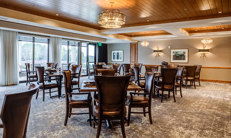 A spacious dining room with multiple dark wooden tables and chairs arranged neatly. Each table is set with glassware and napkins. Large windows with sheer white curtains allow natural light to fill the room, and there are elegant chandeliers hanging from a wooden ceiling. The walls are decorated with framed artwork and sconces.