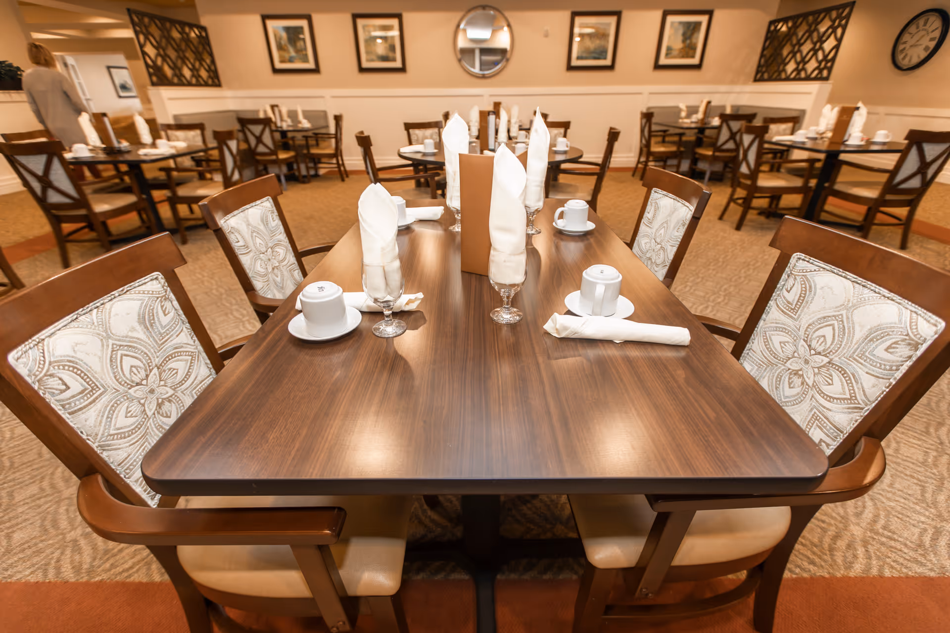 A dining room in a senior living facility with wooden tables and chairs. The tables are set with white napkins folded in glasses, white cups, and saucers. The room has framed artwork on the walls, a round mirror, and a clock. The carpet is patterned and the lighting is warm.