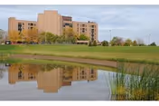 A multi-story beige building reflected in a calm pond with green grass and trees surrounding the area under a cloudy sky.