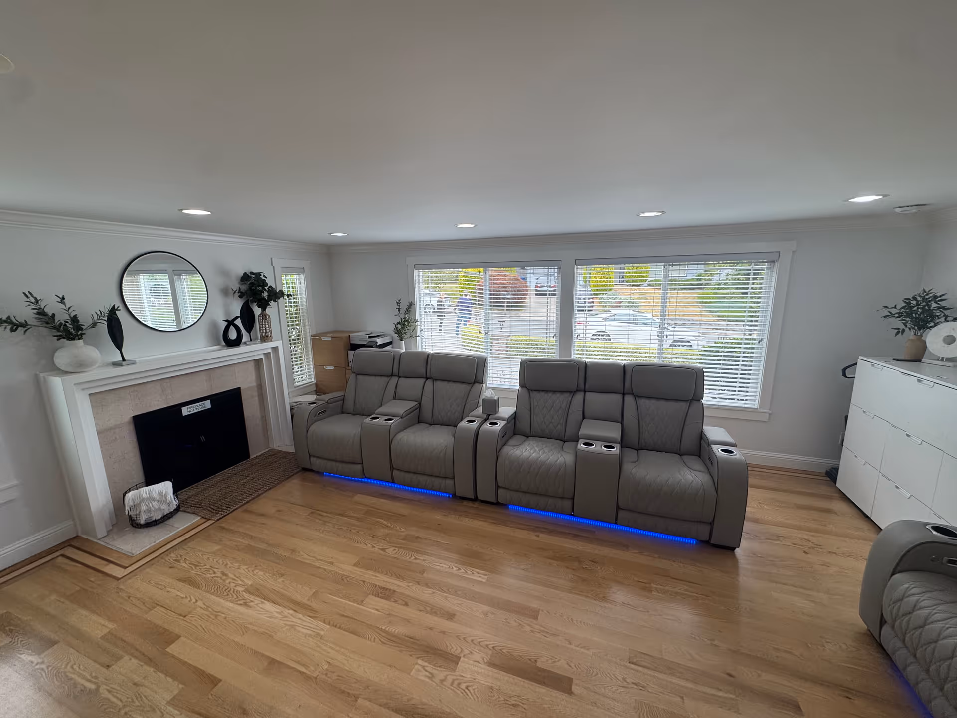 A bright living room with light wooden flooring and white walls. There are two rows of gray reclining chairs with blue LED lights underneath, positioned in front of large windows with white blinds. To the left, there is a white fireplace with decorative vases and a round mirror above it. On the right side, there is a white cabinet with plants and decor items on top.
