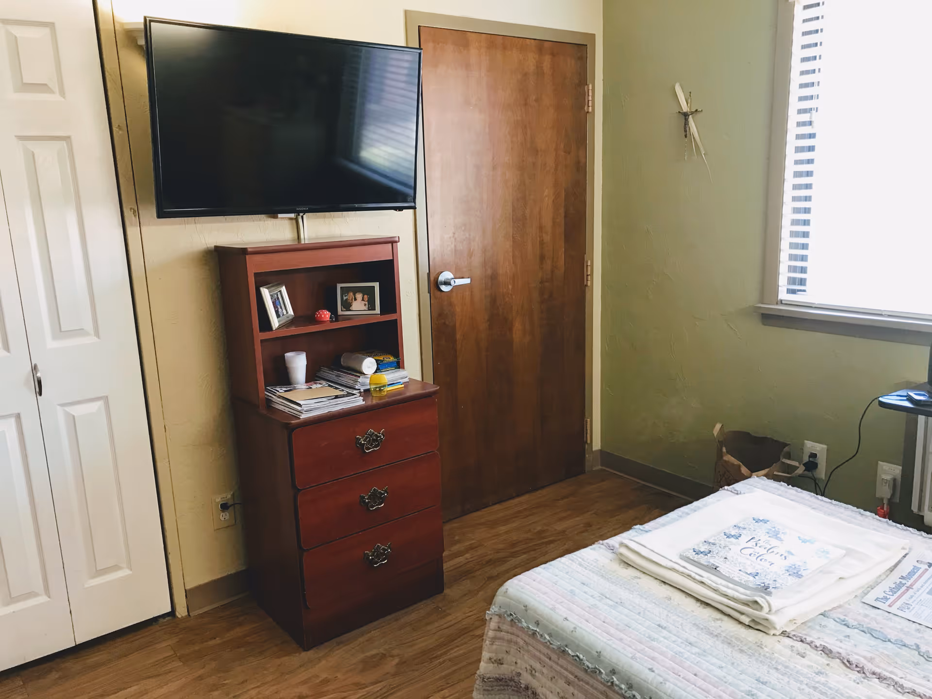 Bedroom with a wall-mounted TV above a wooden dresser, a closed wooden door, a window with blinds, and a bed with folded linens.