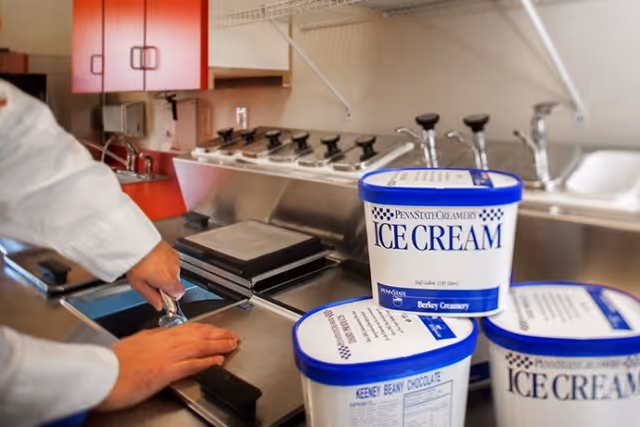 Staff member working at a stainless-steel serving station with tubs of Penn State Creamery ice cream in the foreground.