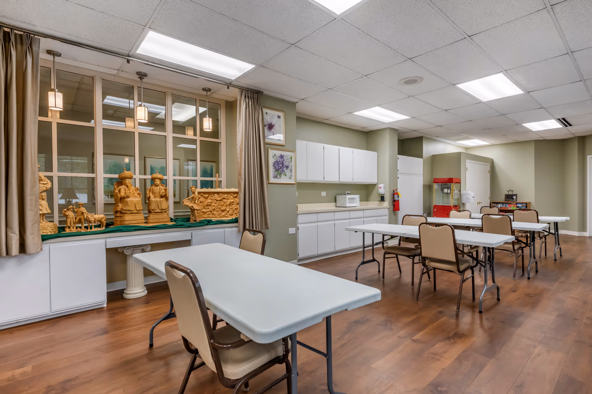 A spacious room with several white folding tables and beige chairs arranged in rows on a wooden floor. The walls are painted light green, and there are white cabinets and a microwave along one wall. Decorative wooden sculptures are displayed on a ledge beneath a large window with beige curtains. The ceiling has recessed fluorescent lighting.
