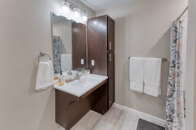 A clean and modern bathroom featuring a white sink with a dark wood vanity and matching tall cabinet. Above the sink is a large mirror with three light fixtures. White towels hang on a ring and a bar, and a floral shower curtain is partially visible on the right side.