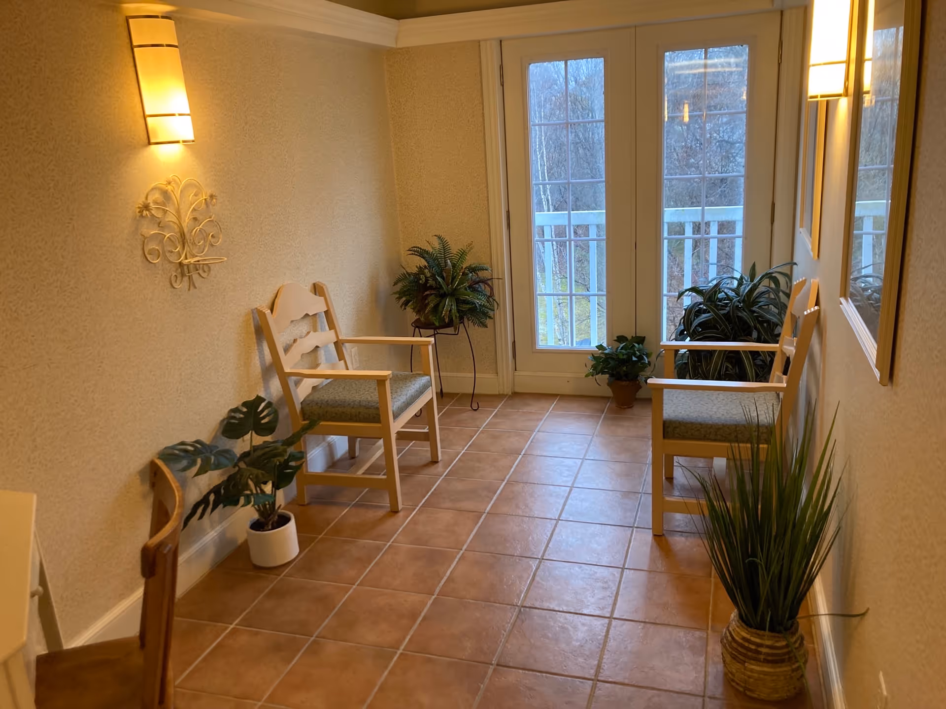 A small sitting area with two wooden chairs with green cushions placed against opposite walls. The floor is tiled with light brown tiles. There are several potted plants placed around the room, including near the chairs and by the glass doors. The walls are light beige with a decorative wall sconce light fixture and a metal wall decoration. The glass doors lead to an outdoor area with a white railing and trees visible outside.