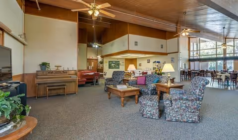 Spacious senior living facility common area with high wooden ceilings, ceiling fans, large windows letting in natural light, floral upholstered armchairs, wooden coffee tables, a piano against the wall, and a dining area with tables and chairs in the background.