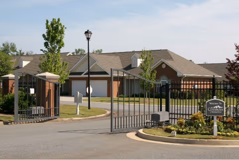 Entrance gate to The Cottages of Milledgeville senior living facility with a black metal fence and gate, a street lamp, landscaped greenery, and brick buildings with garages in the background under a clear sky.
