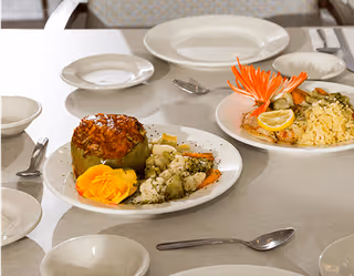 A dining table set with plates and utensils, featuring two plates of food. One plate has a stuffed green bell pepper with a yellow garnish and mixed vegetables, while the other plate has rice, lemon slices, and a decorative orange garnish.