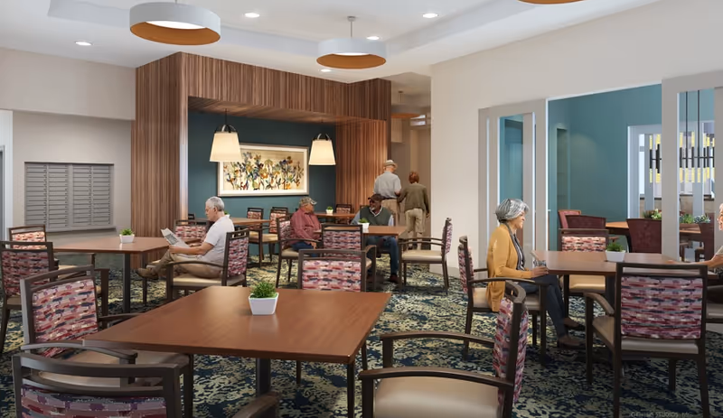 A senior living community dining area with several elderly residents seated at wooden tables with patterned chairs. The room features a blue accent wall with a floral painting, pendant lights, and a carpet with a floral design. There are small potted plants on the tables and a mail area with mailboxes on the left side.