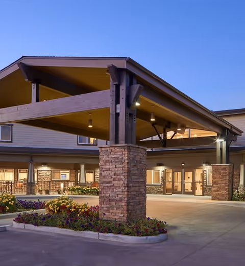 Entrance of Ralston Creek Neighborhood Assisted Living and Memory Care facility during dusk, featuring a covered drop-off area with wooden beams and stone pillars, surrounded by flower beds and well-lit building exterior.