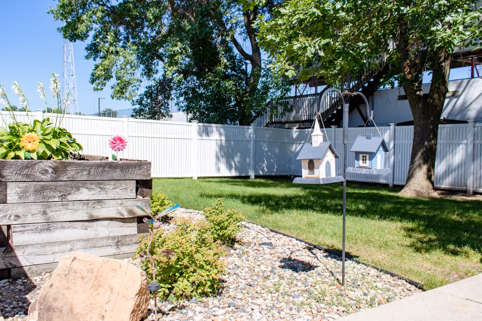 Outdoor garden area with a wooden raised planter box containing flowers, a small rock garden with green plants, two decorative birdhouses hanging from a metal pole, a large tree, and a white fence in the background under a clear blue sky.