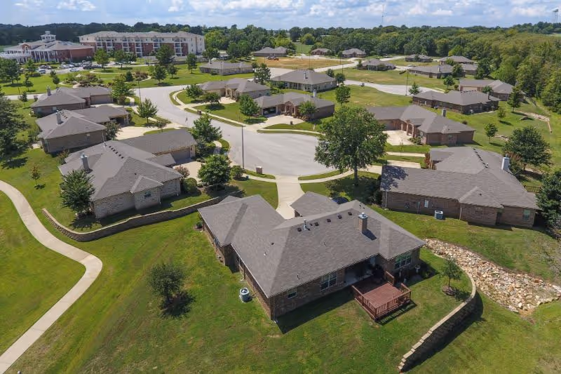 Aerial view of Meadow Lake Senior Living showing multiple single-story residential buildings with gray roofs arranged around curved roads and green lawns, with trees and sidewalks throughout the community.