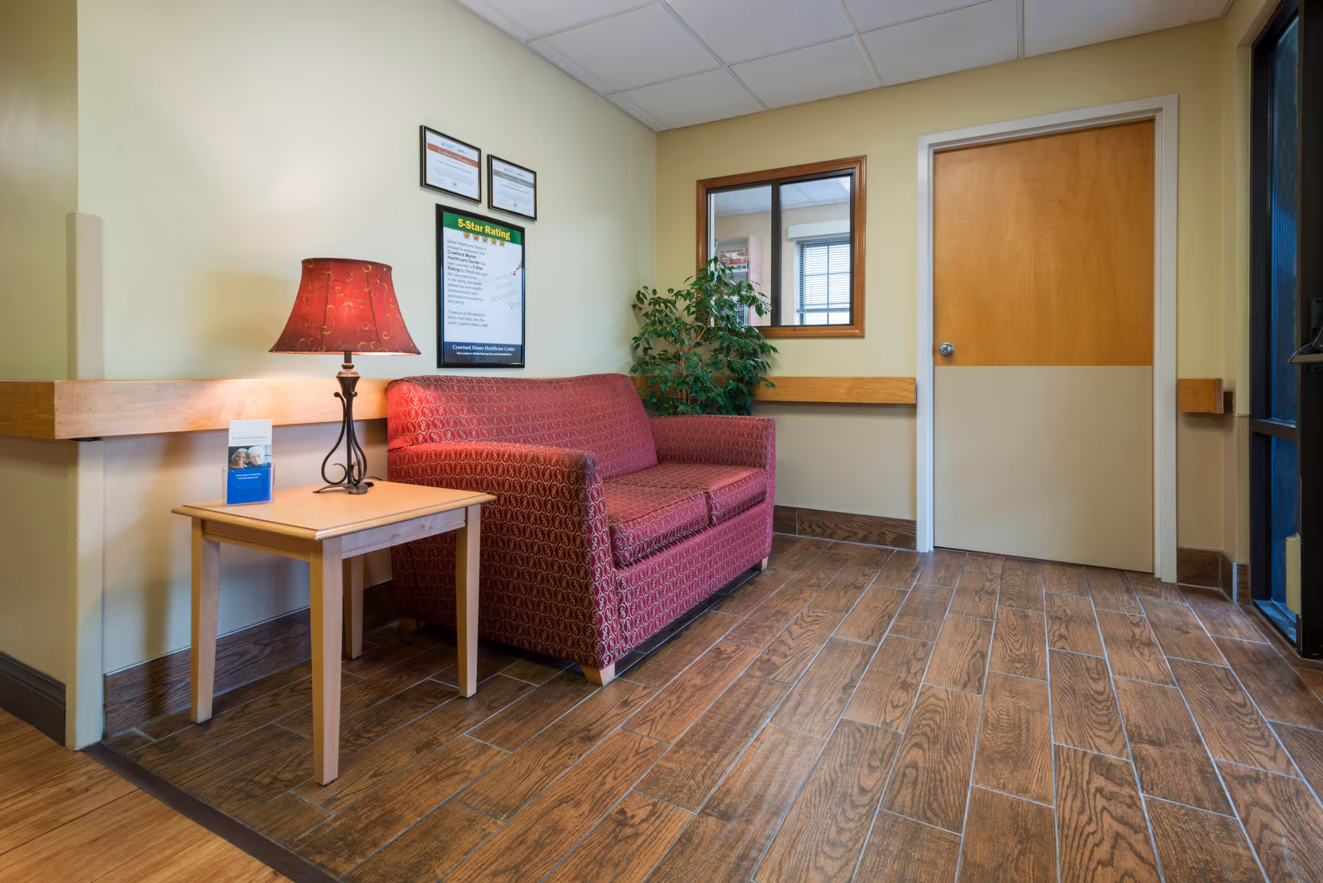 A small waiting area inside Crawford Manor Health Care Center featuring a red patterned loveseat, a wooden side table with a red lamp and a brochure, a green potted plant, a wooden door, and a window looking into another room. The walls are painted light yellow and the floor has wood-like tiles.
