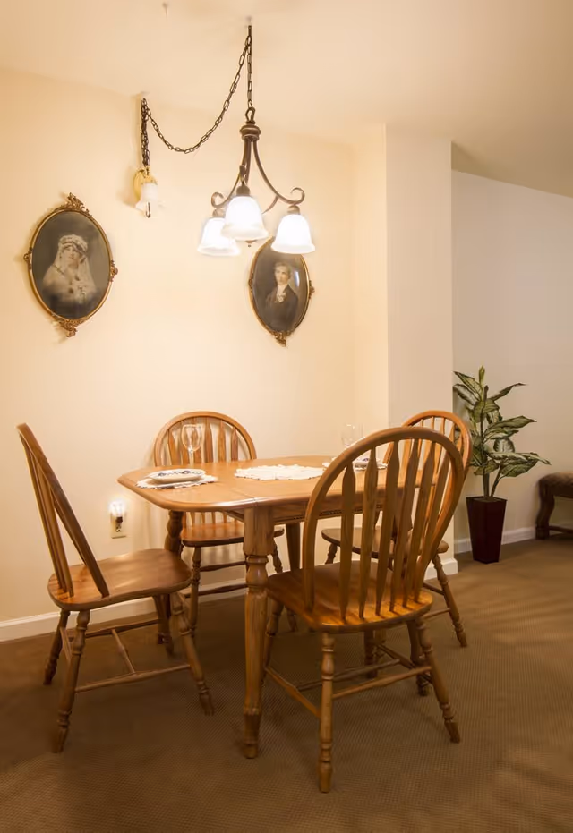 A small dining area with a wooden table and four wooden chairs. The table is set with placemats and wine glasses. Above the table hangs a chandelier with four lights. Two oval framed portraits are mounted on the wall behind the table. A potted plant is placed in the corner of the room.