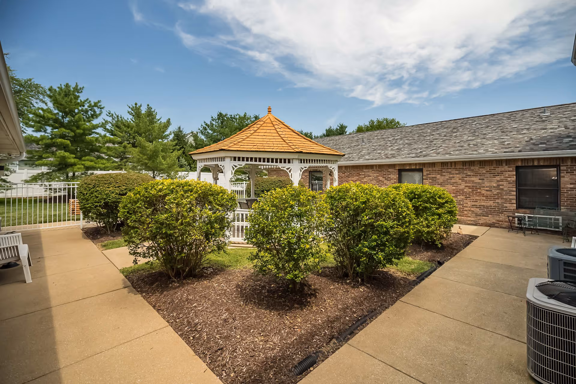 Courtyard with a white gazebo surrounded by trimmed shrubs, concrete walkways and a brick building under a partly cloudy sky.