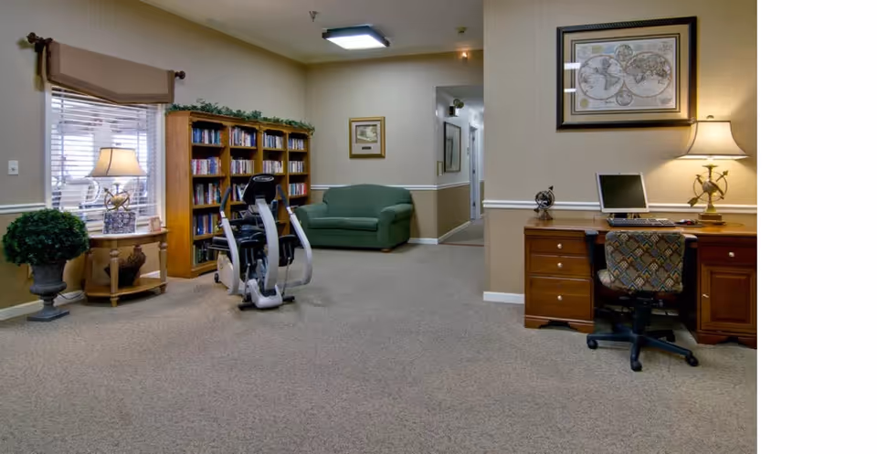 Interior room with beige walls and carpeted floor featuring a wooden desk with a computer and lamp, a green armchair, a bookshelf filled with books, an exercise bike, a small round table with a lamp, and framed artwork on the walls.