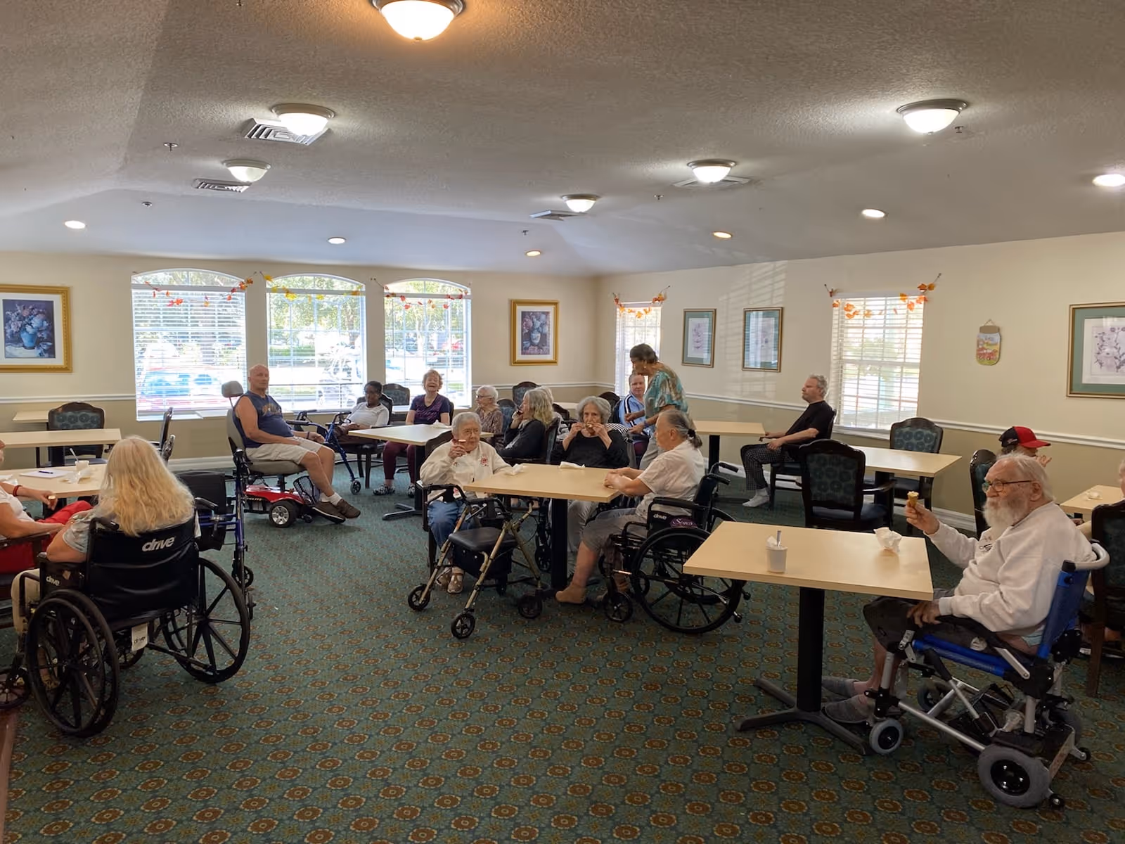A group of elderly people, some in wheelchairs and others seated, gathered in a well-lit common room with tables and chairs. The room has large windows with blinds, framed artwork on the walls, and a patterned carpet. A caregiver is interacting with some of the residents.