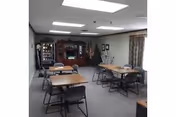 Communal dining/activity room with several rectangular tables and folding chairs, a wooden hutch and vending machine along the back wall, and ceiling lights.