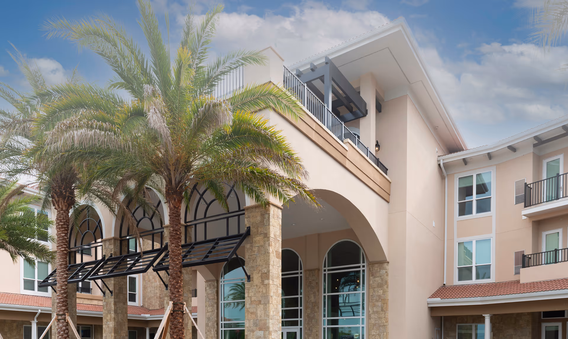 Exterior view of Heartis Venice Assisted Living facility showing a multi-story building with large arched windows, stone pillars, balconies, and palm trees in front under a partly cloudy sky.