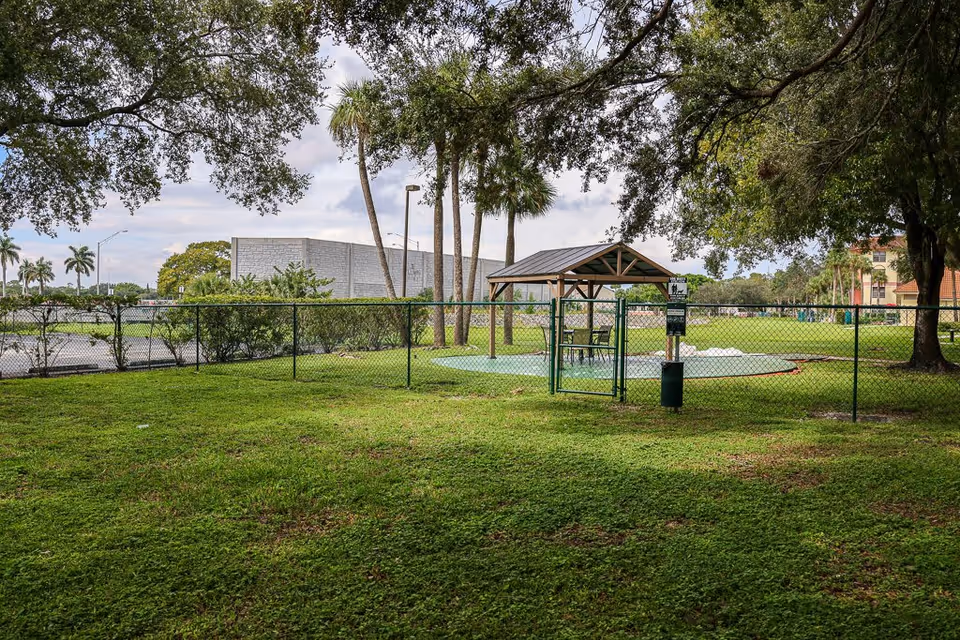 A fenced outdoor area with a small covered pavilion containing a table and chairs, surrounded by green grass and trees. Palm trees and other greenery are visible in the background under a partly cloudy sky.