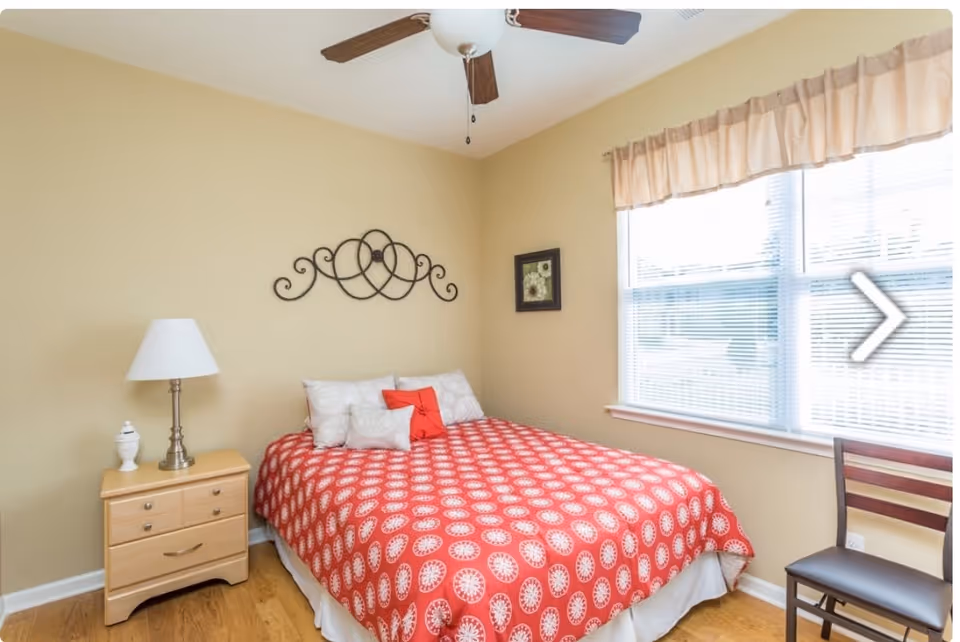 A bedroom with a bed covered in a red patterned bedspread and several pillows. There is a wooden nightstand with a lamp and decorative item on it to the left of the bed. A decorative metal wall art piece hangs above the bed, and a framed picture is on the adjacent wall. A window with blinds and a valance is on the right side, next to a wooden chair. The room has light beige walls and a ceiling fan with wooden blades.