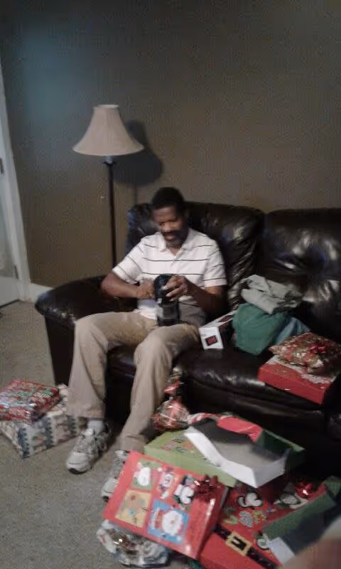 A man sitting on a dark brown leather couch in a living room, holding a black object. There are several wrapped Christmas presents and gift boxes on the floor and couch around him. A floor lamp with a beige lampshade is positioned behind the couch against a plain wall.