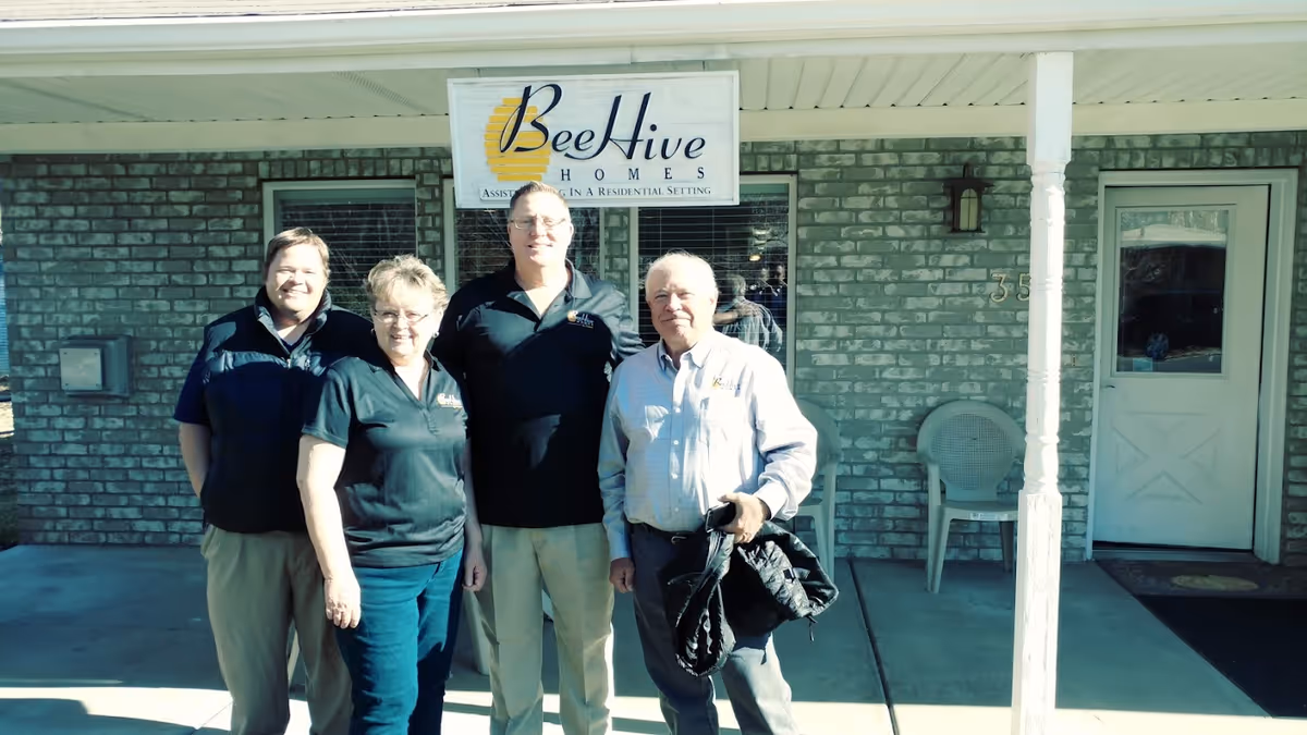 Four adults standing outside the entrance of Beehive Homes of Logan, a brick building with a white door and a sign above the group that reads 'Beehive Homes Assisted Living in a Residential Setting'.