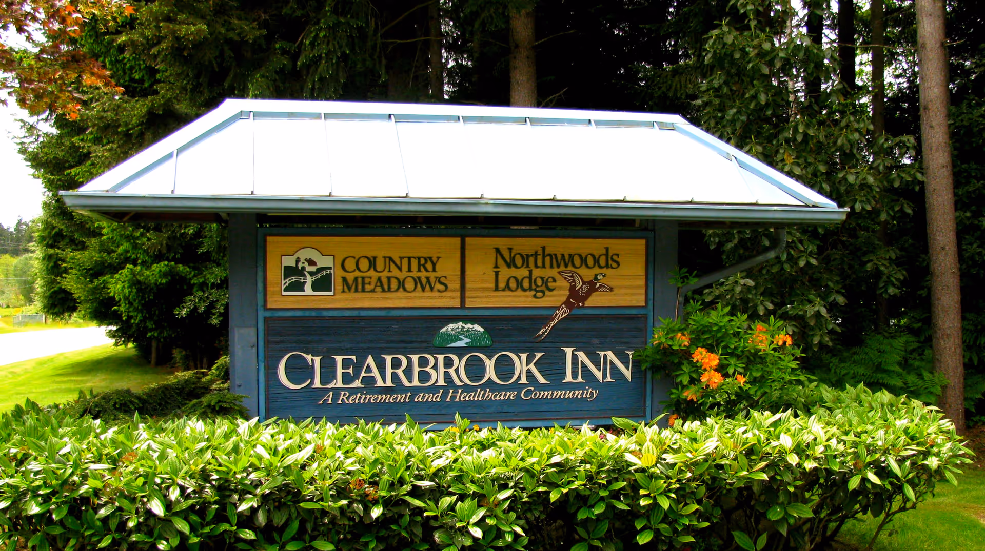 Outdoor sign for Clearbrook Inn, a retirement and healthcare community, with logos for Country Meadows and Northwoods Lodge. The sign is surrounded by green bushes and trees.