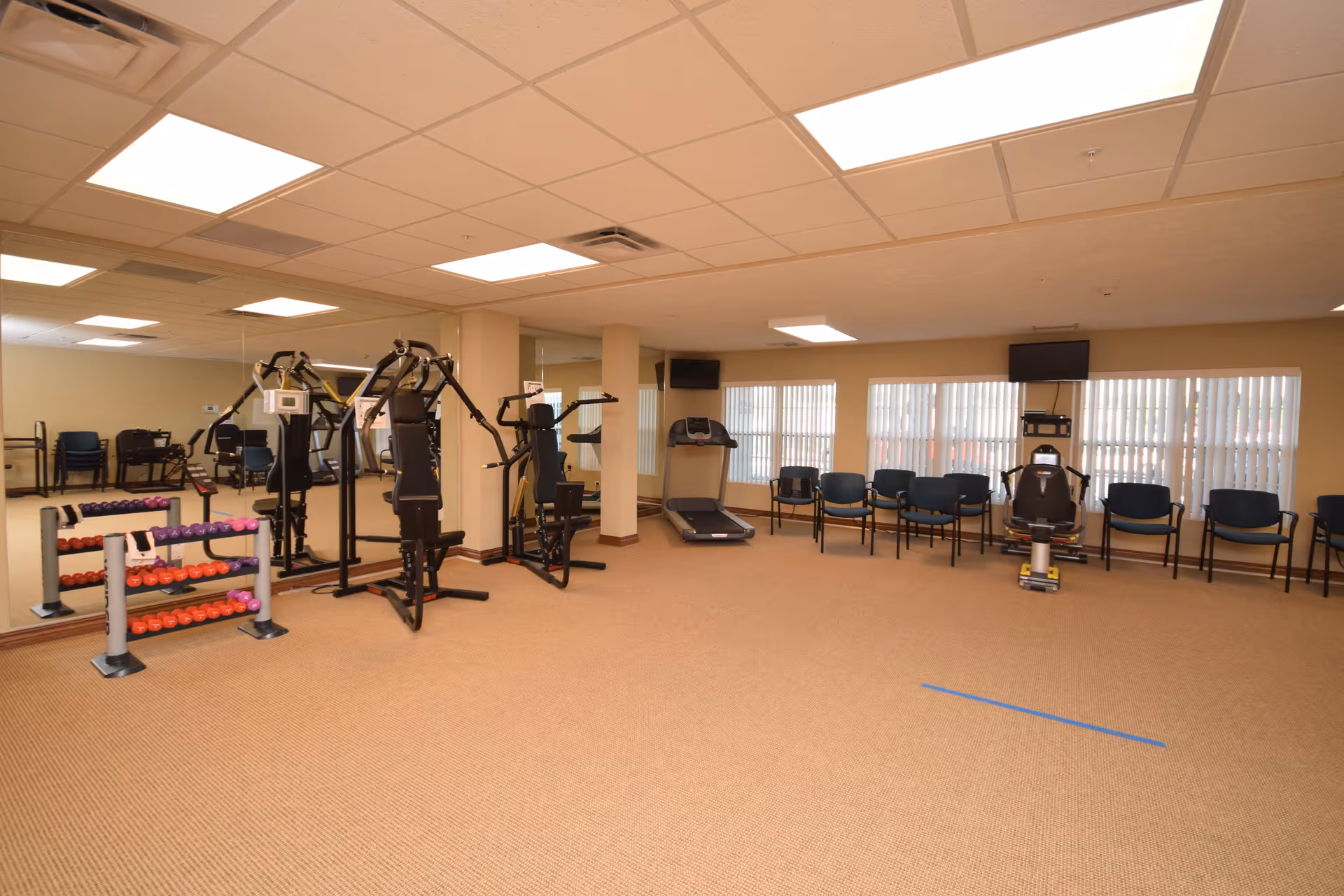 A spacious fitness room with exercise equipment including a multi-station gym machine, a treadmill, and a recumbent bike. There are several chairs lined up against the wall with large windows covered by vertical blinds. The room has beige walls, a carpeted floor, and a drop ceiling with fluorescent lighting.
