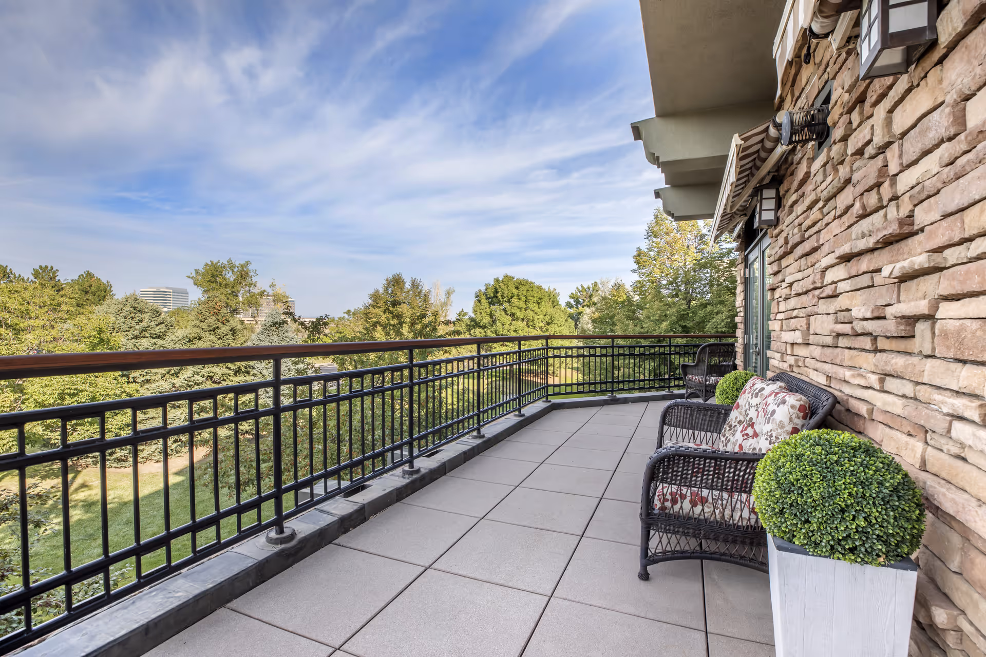 A spacious outdoor balcony with a black metal railing overlooking green trees and a clear blue sky. The balcony has a tiled floor and is furnished with black wicker chairs with floral cushions and a white planter with a green shrub. The building wall is made of stone with mounted lights and an awning above the door.