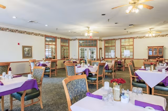 A dining room in a senior living facility with multiple tables covered in white and purple tablecloths. Each table is set with glasses, cups, napkins, and small flower arrangements. The room has carpeted floors, wooden chairs with patterned upholstery, ceiling fans with lights, and windows with wooden frames. There is a cabinet and a grandfather clock against the walls.
