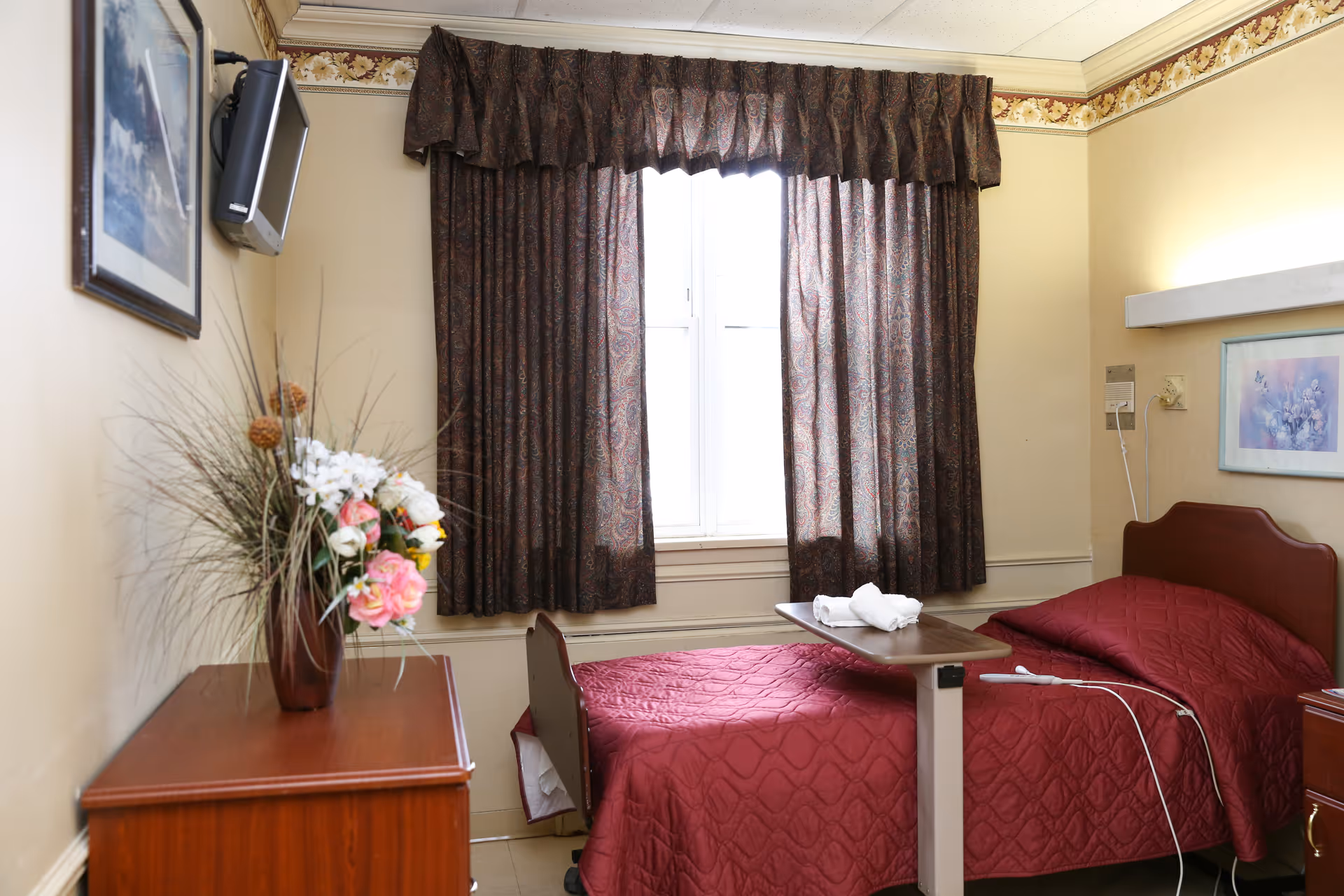 A small bedroom in a senior living facility with a single bed covered in a red quilted bedspread. There is a wooden bedside table with a floral arrangement in a vase. A window with dark patterned curtains is centered on the back wall. A small flat-screen TV is mounted on the left wall above a framed picture. A hospital-style overbed table is positioned over the bed with a folded towel on it.