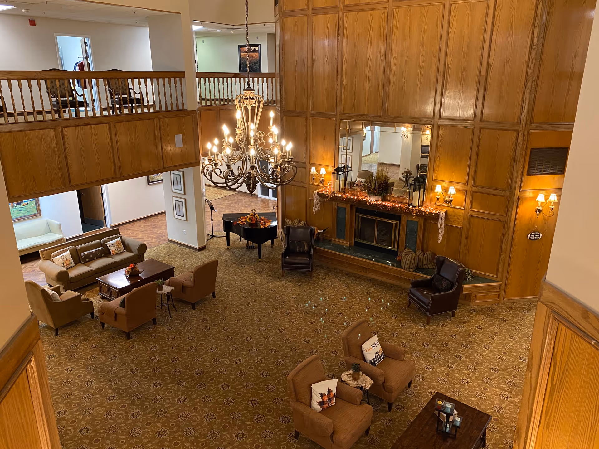 A spacious living room area with a large chandelier hanging from the ceiling. The room features a fireplace with a wooden paneled wall and a large mirror above it. There are several seating arrangements including sofas, armchairs, and small tables. A grand piano is positioned near the back wall. The carpet has a patterned design and the overall decor has a warm, inviting feel.