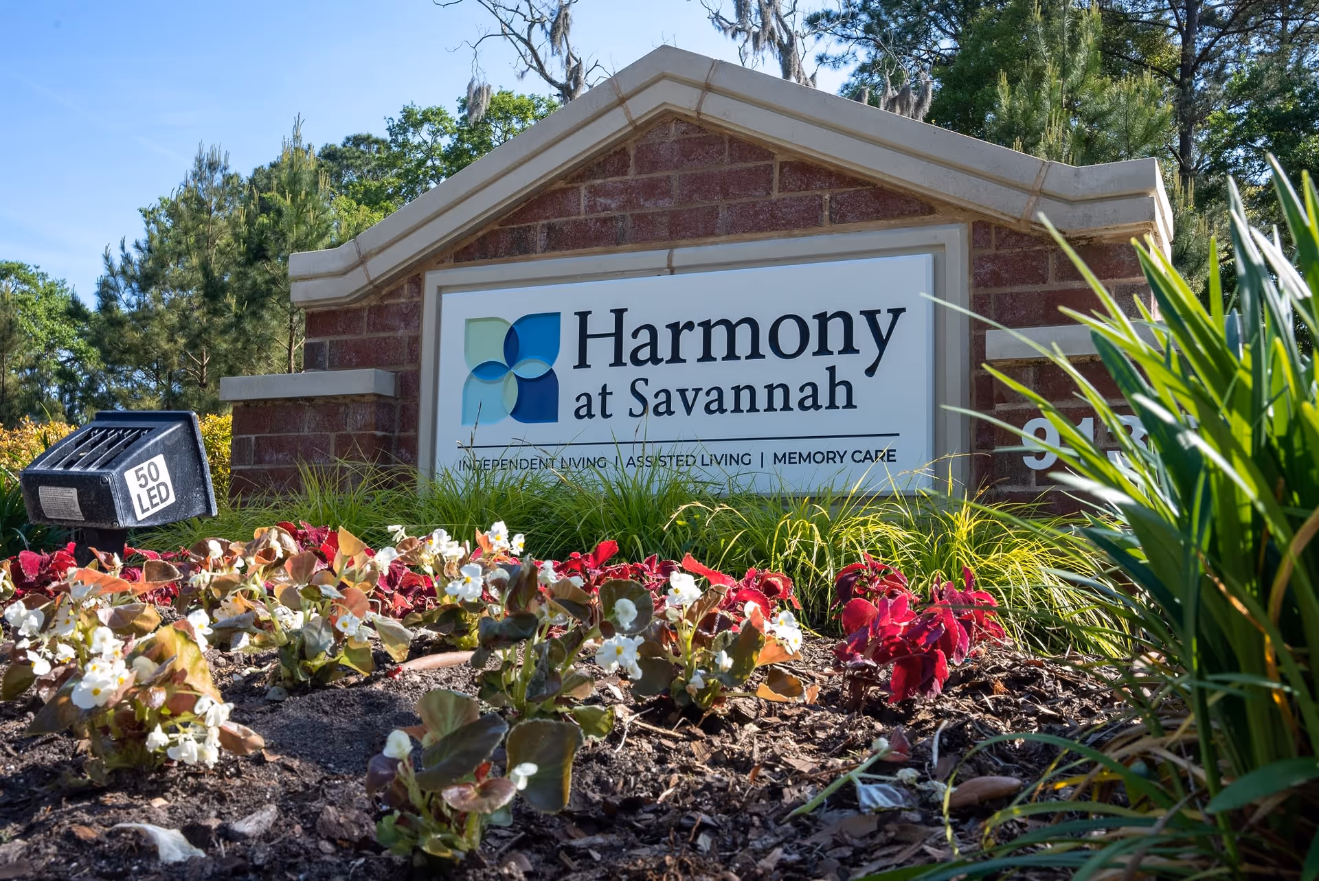 A brick sign for Harmony at Savannah surrounded by flowers and greenery, with trees and a clear sky in the background.