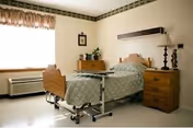 A hospital-style bedroom with a single bed featuring a wooden headboard and footboard, covered with a patterned bedspread. Next to the bed is a wooden nightstand with a lamp and decorative items. A window with curtains allows natural light into the room, and there is an air conditioning unit below the window. The walls are light-colored with a decorative border near the ceiling.