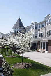 Exterior view of Munhall Retirement Residence showing a multi-story building with large windows and a turret. In the foreground, there are blooming white flowering trees, a green lawn, a paved walkway, and a wooden bench under a clear blue sky.