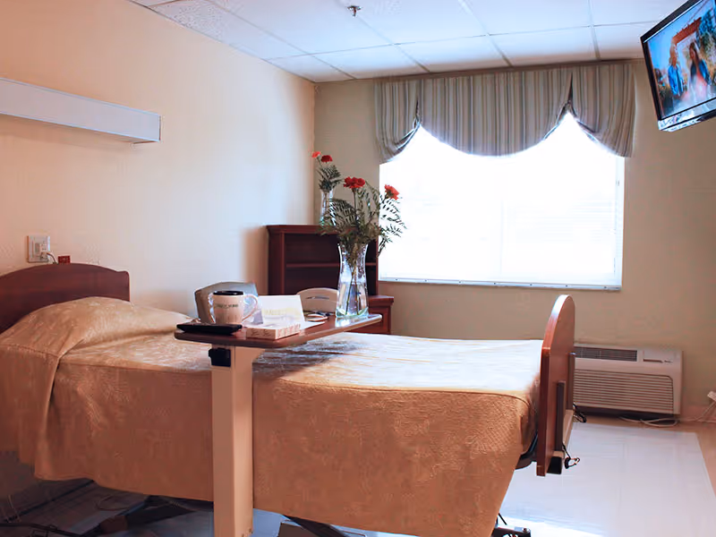 A neatly arranged patient bedroom with a hospital-style bed, overbed table holding flowers and items, a curtained window and a wall-mounted TV.