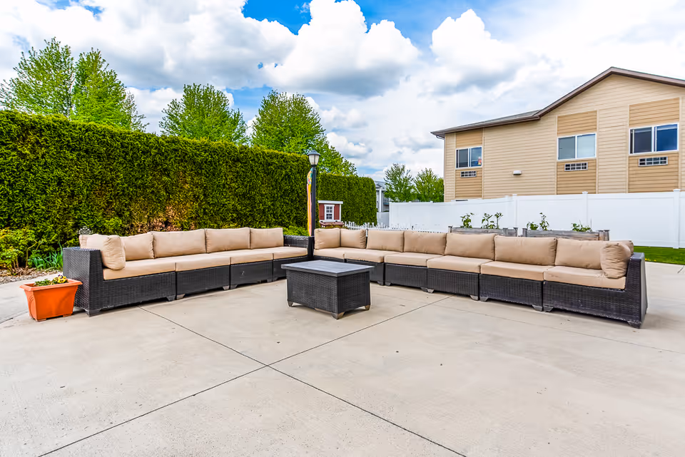 Outdoor courtyard with a large L-shaped wicker sectional sofa and central coffee table on a concrete patio, with a hedge and building in the background.