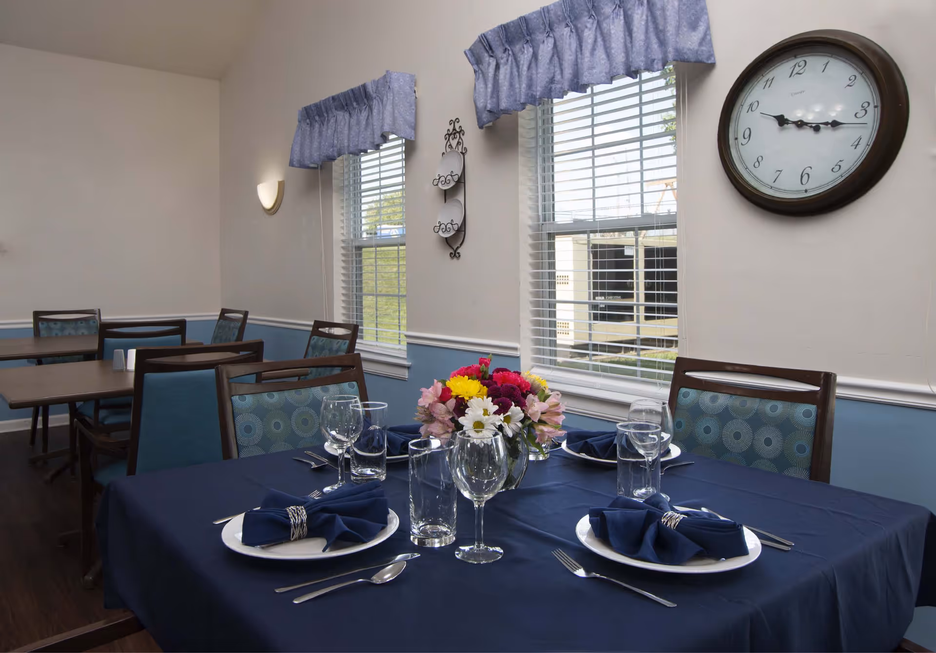 Dining room with tables and chairs, one table set with plates, silverware, glasses, navy blue napkins, and a centerpiece of colorful flowers. Two windows with blue valances and white blinds let in natural light. A round wall clock shows the time as 3:15.
