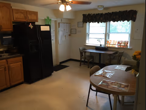 Interior view of a kitchen and dining area with wooden cabinets, a black refrigerator, a ceiling fan with lights, two tables with chairs, windows with blinds and a valance, and various papers and decorations on the walls and tables.