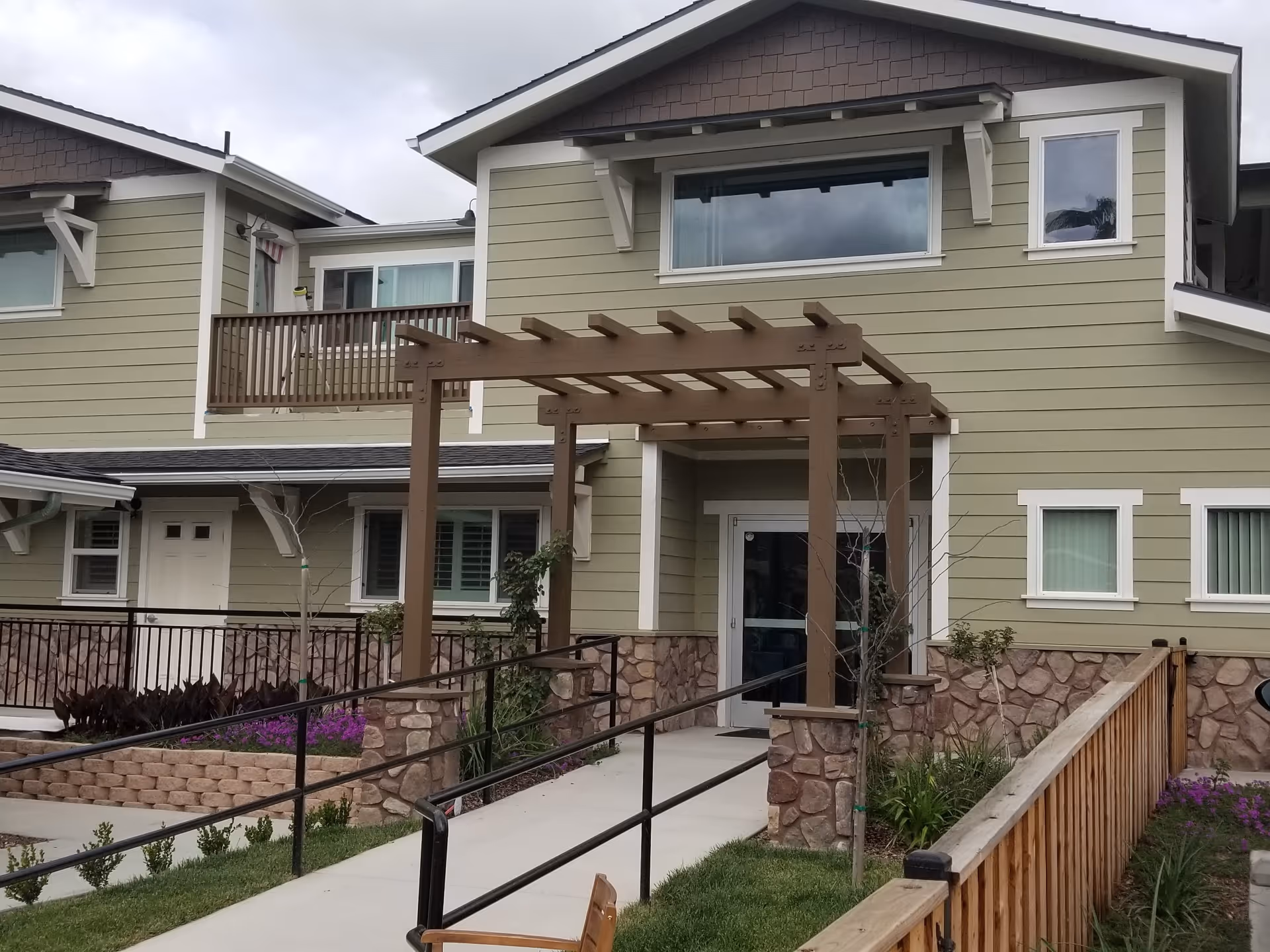 Front entrance of a two-story building with a wooden pergola, wheelchair ramp, and stone-faced lower walls.