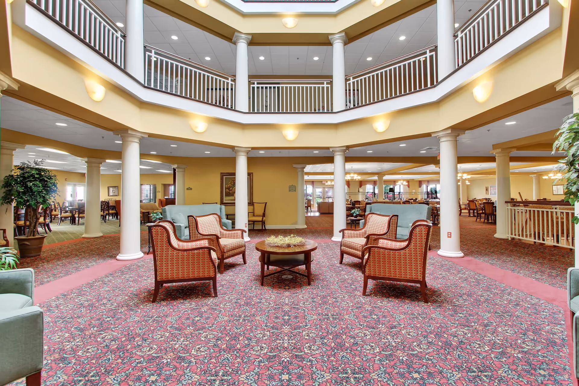 Spacious and well-lit common area in a senior living facility with patterned red carpet, several armchairs and sofas arranged around a round wooden coffee table, white columns supporting an upper balcony, and warm yellow walls.