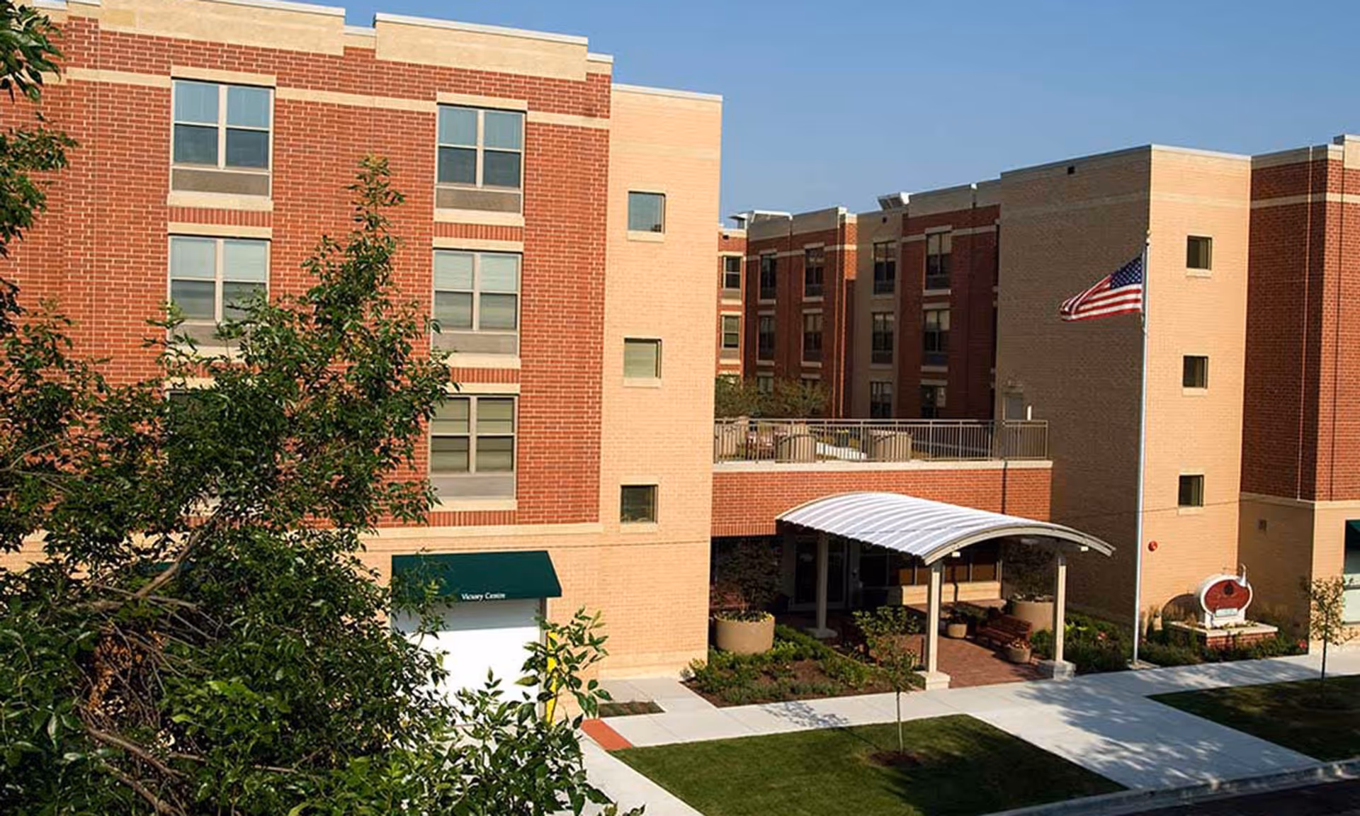 Exterior view of a multi-story brick and beige building with multiple windows, an American flag on a pole, a covered entrance with a white canopy, and landscaped greenery including trees and shrubs.
